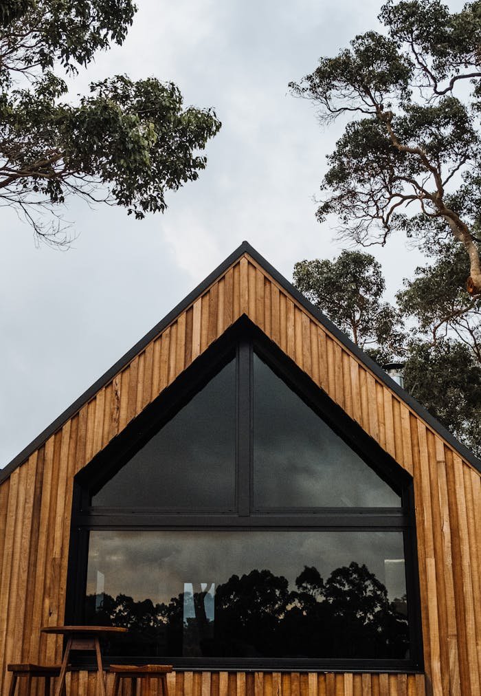 About A stylish wooden cabin featuring a large triangular glass window, surrounded by trees.